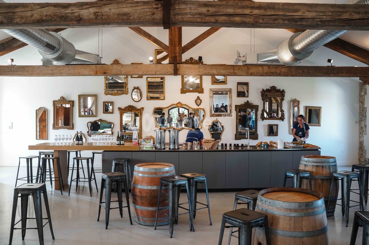 Salle de dégustation avec bar en bois, tonneaux, tabourets et murs décorés de miroirs anciens.
