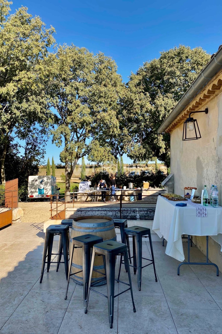 Terrasse ensoleillée avec tables hautes, tabourets, table dressée et vue sur un jardin arboré en arrière-plan.