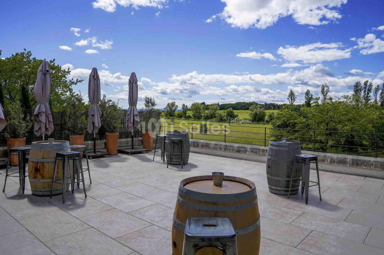 Terrasse ouverte avec tables tonneau, chaises hautes et parasols fermés - vue sur paysage verdoyant et ciel dégagé.