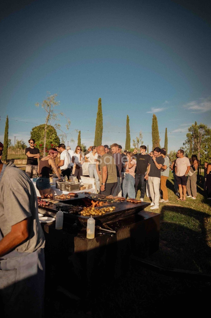 Groupe de convives debout autour d'un barbecue en plein air, cyprès en arrière-plan, ciel dégagé.