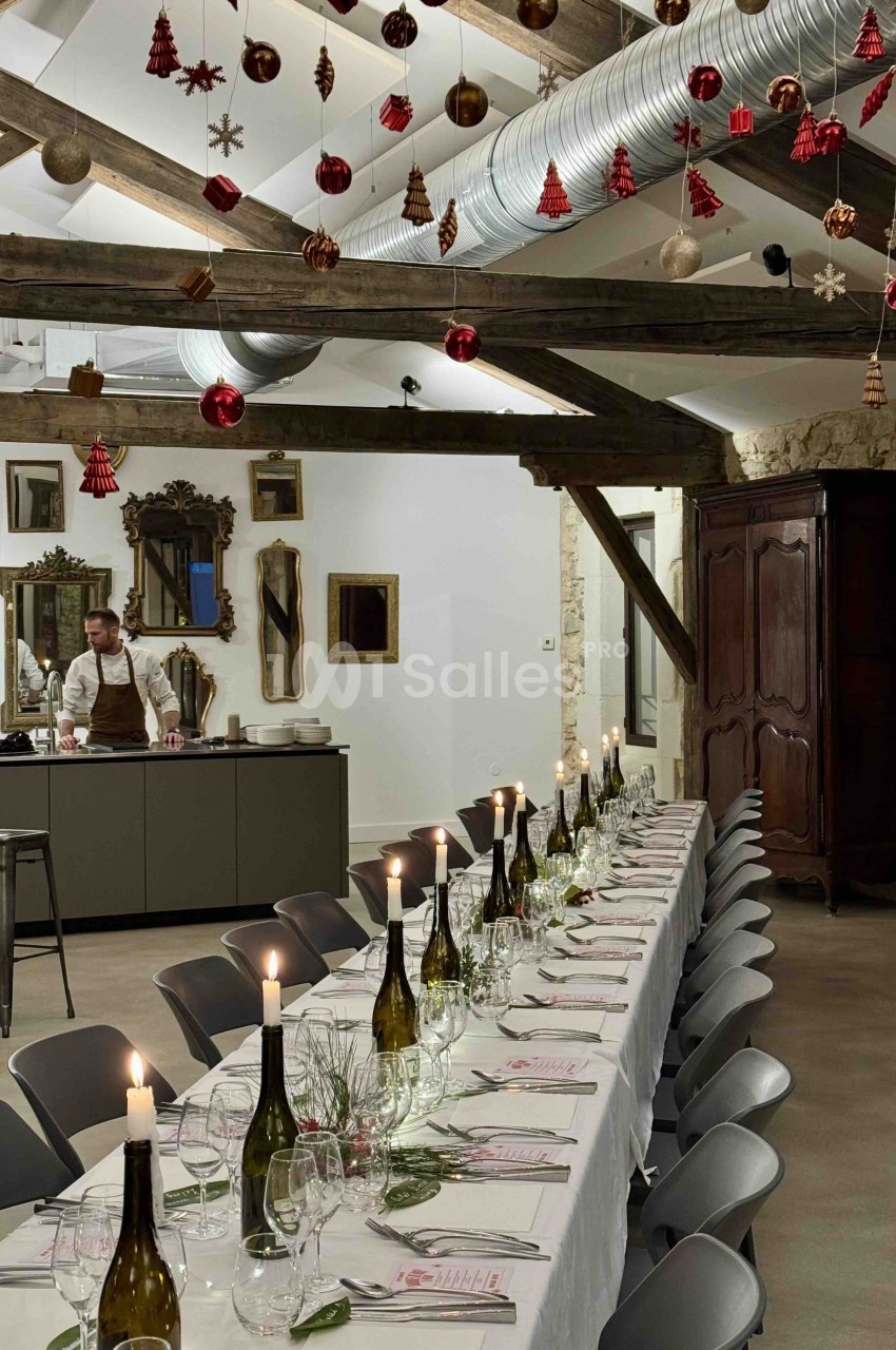 Salle en pierre avec poutres apparentes, table dressée pour environ 20 convives, décoration hivernale suspendue.
