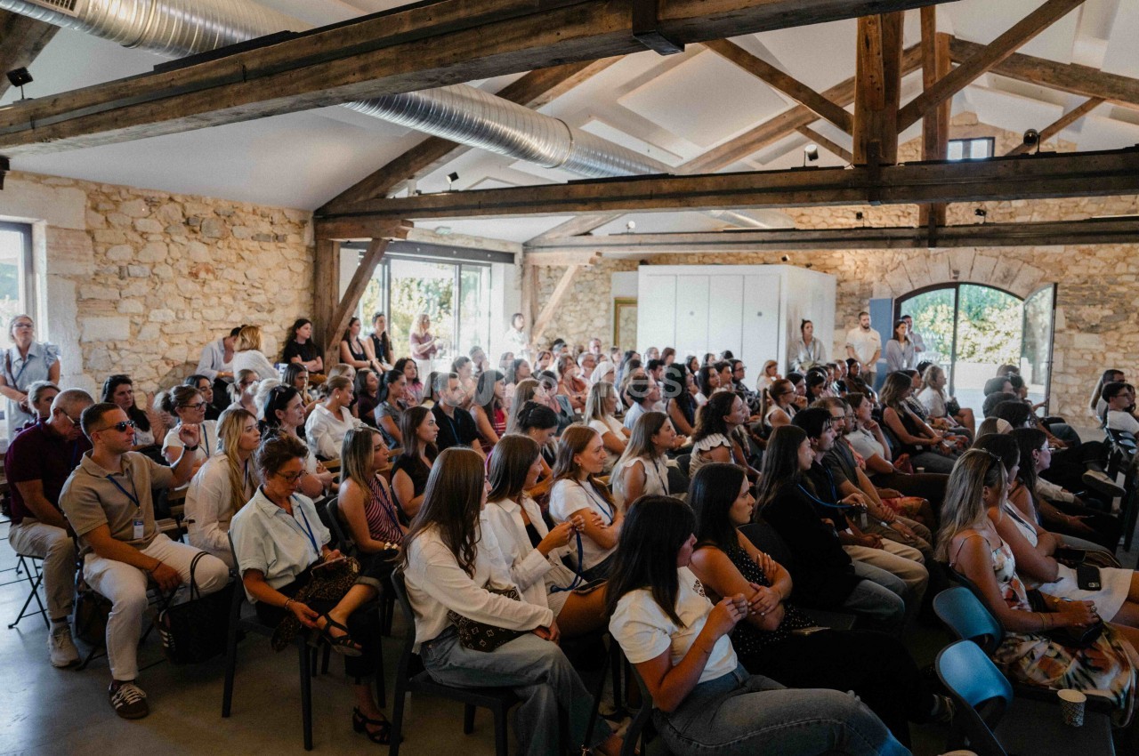 Salle intérieure aux murs en pierre et poutres apparentes, remplie de nombreux convives assis en configuration théâtre.