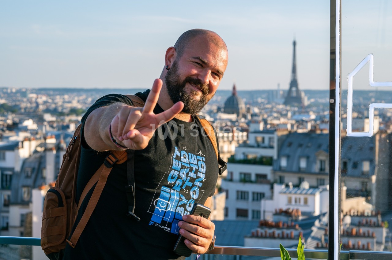 Un homme souriant fait un signe de paix devant une vue panoramique de Paris avec la tour Eiffel en arrière-plan.