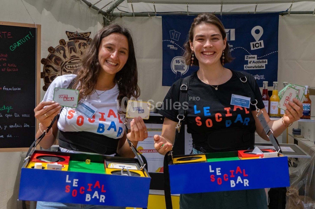 Deux femmes souriantes présentent des jeux et accessoires dans un stand extérieur sous une tente.