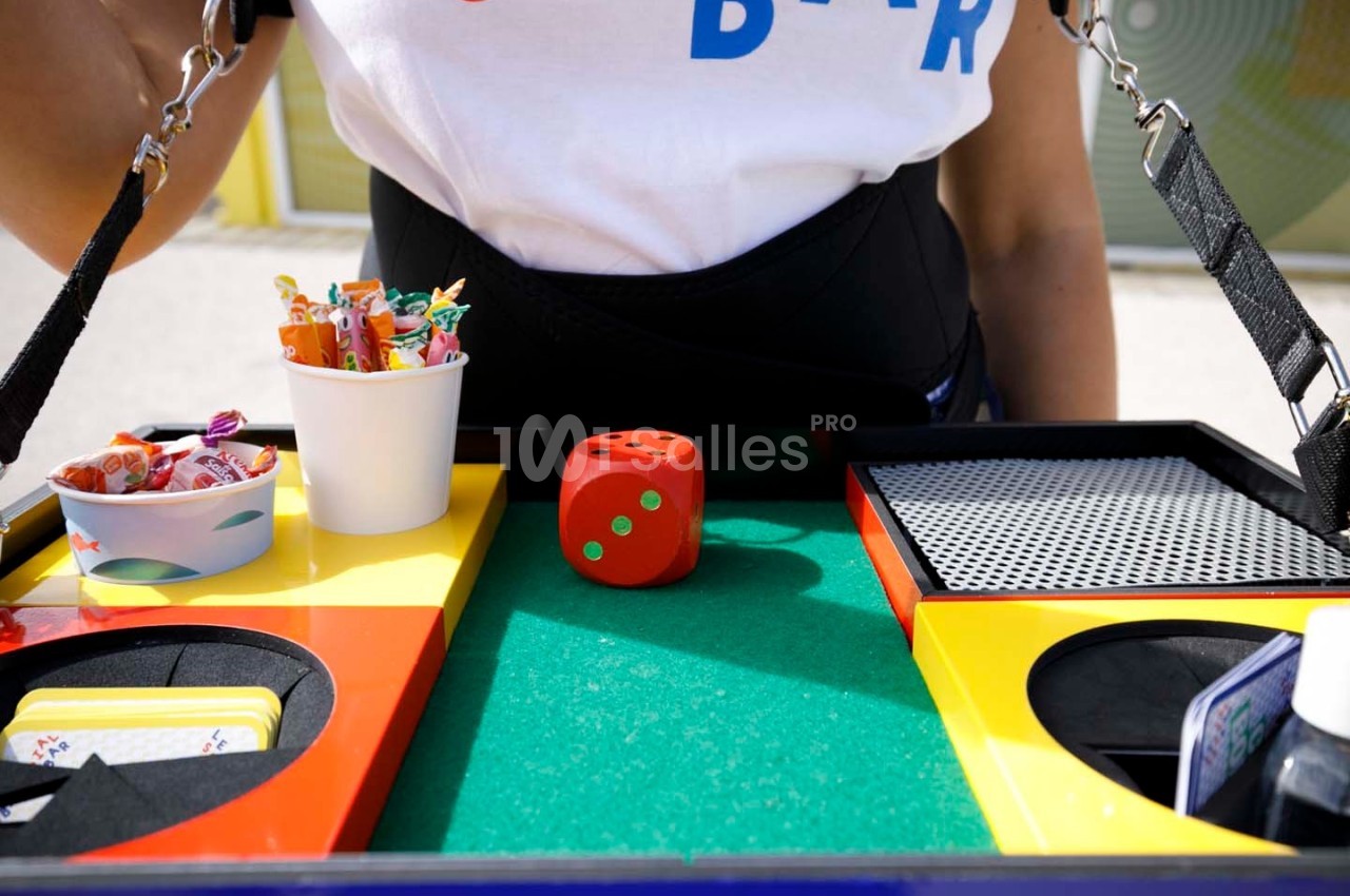 Plateau coloré contenant un dé rouge, des cartes, des bonbons et divers accessoires, tenu par une personne.
