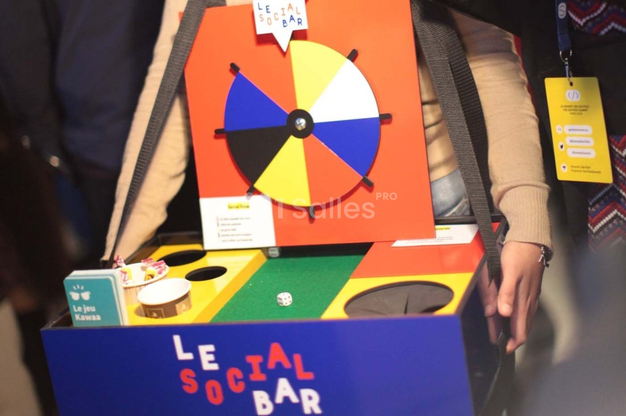 Une personne tient un jeu coloré avec une roue et des compartiments, marqué ’Le Social Bar’.