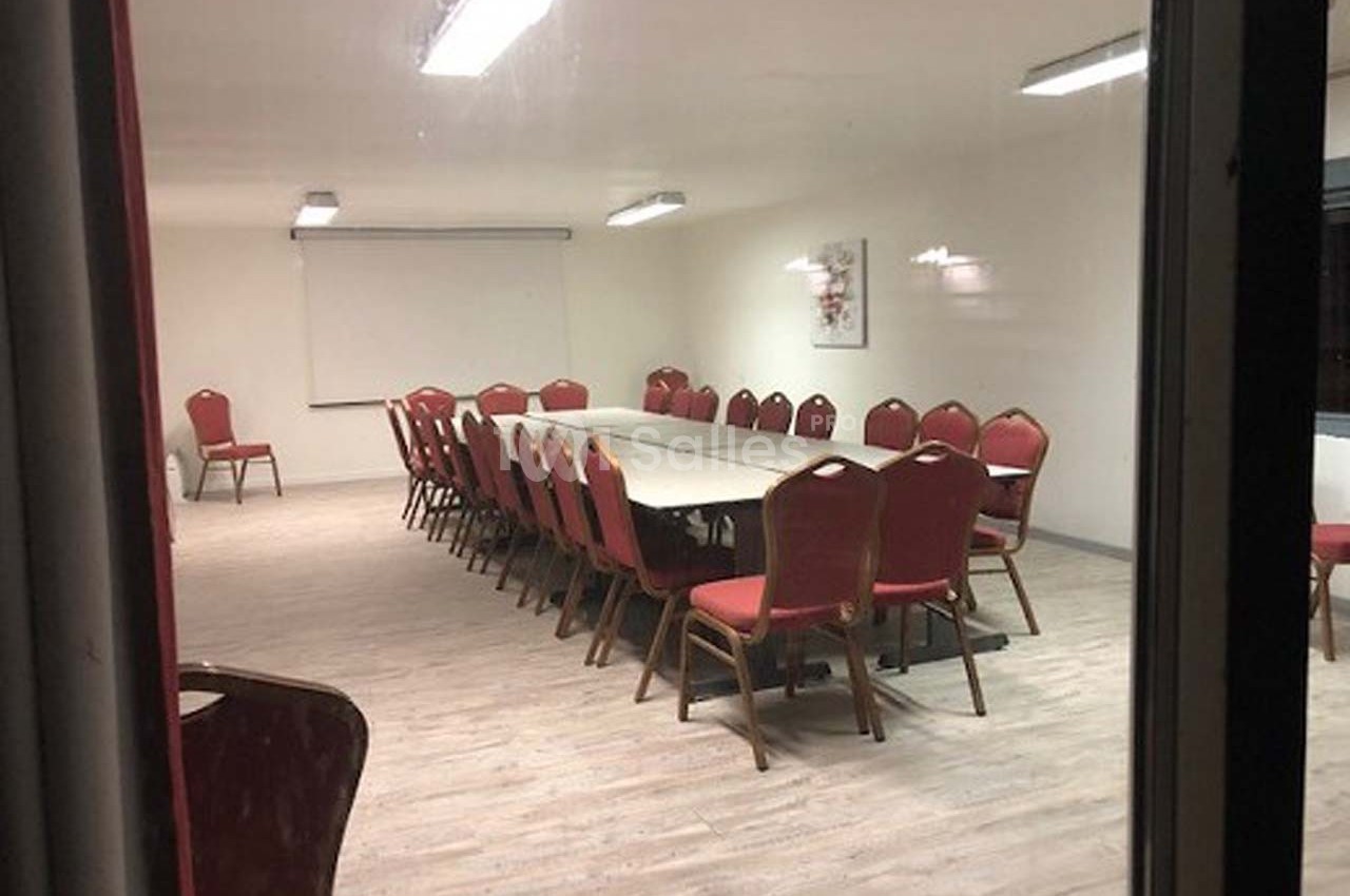 Salle de réunion vide avec une grande table rectangulaire entourée de chaises rouges, éclairée par des néons au plafond.