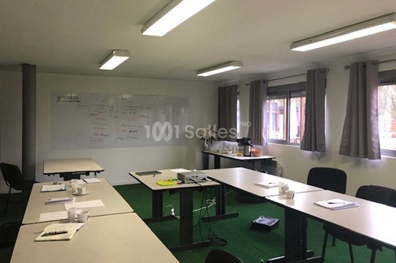 Salle de réunion lumineuse avec tables disposées en U, tableau blanc au mur et matériel de travail sur les tables.
