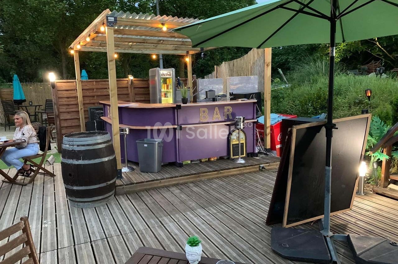 Bar extérieur en bois avec comptoir violet, éclairé par des guirlandes, entouré de tables et parasols sur une terrasse.