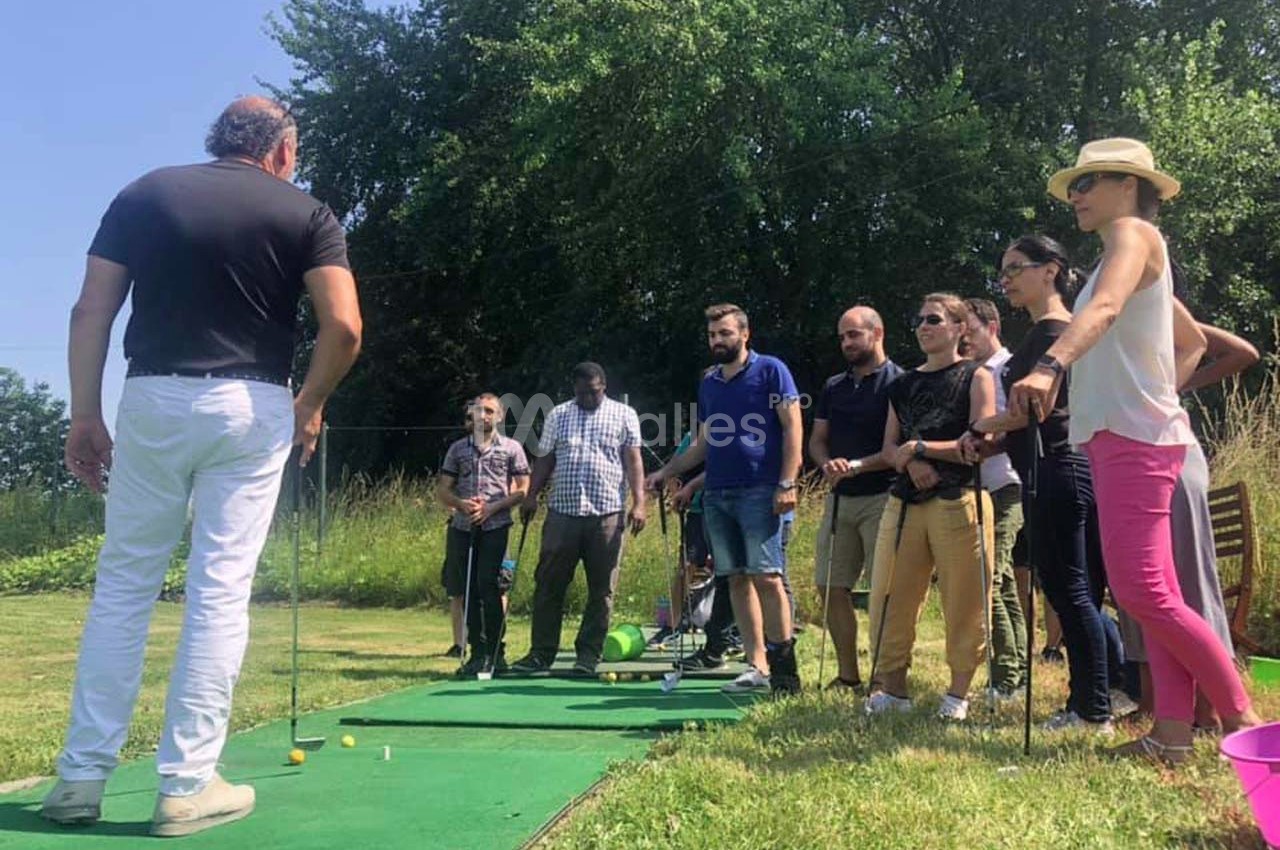 Un groupe de personnes écoute un instructeur de golf sur un terrain en plein air, avec des arbres en arrière-plan.