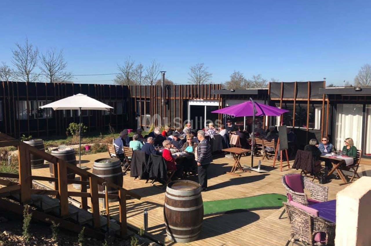 Personnes attablées en terrasse ensoleillée avec parasols colorés, entourées de végétation et de barriques en bois.