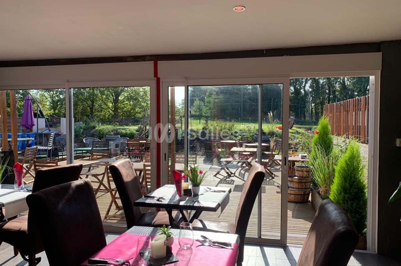 Salle de restaurant lumineuse avec vue sur une terrasse en bois aménagée et un jardin verdoyant à l'extérieur.