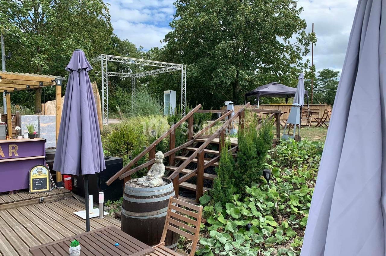 Terrasse extérieure avec tables, parasols, statue décorative et végétation, surplombant un espace verdoyant.