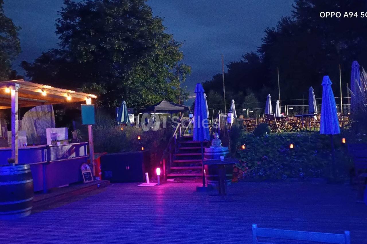 Terrasse extérieure éclairée en soirée avec des parasols, des tables et un bar en bois sous des lumières tamisées.