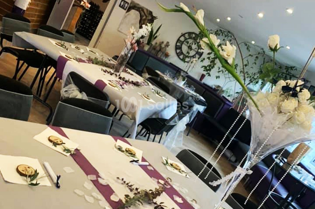 Salle décorée pour un événement avec tables dressées, nappes blanches, chemins de table violets et compositions florales.