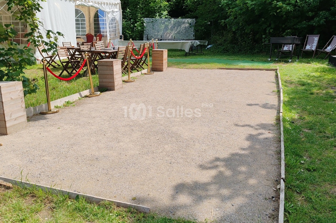 Terrain de pétanque en gravier entouré de verdure, avec des tables et une tente blanche en arrière-plan.
