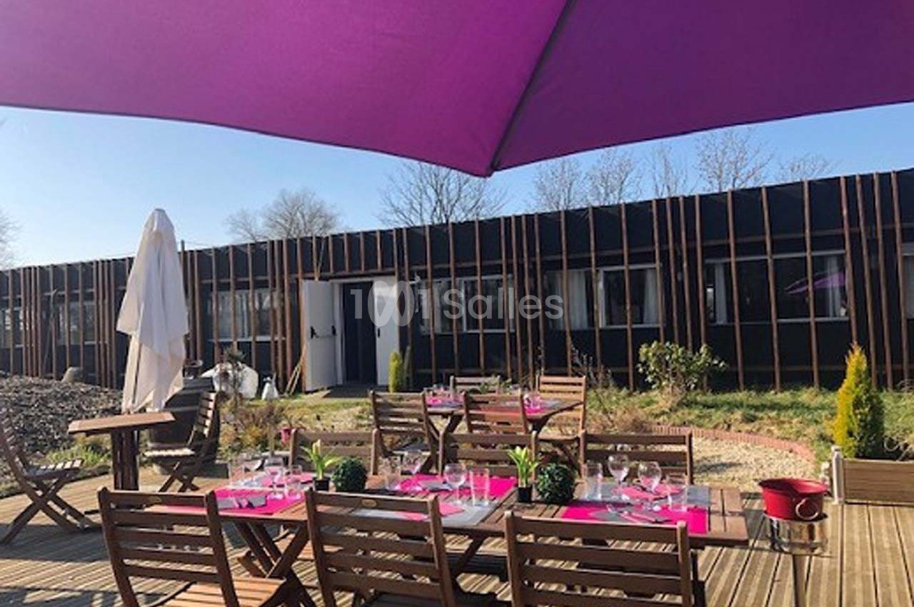 Terrasse en bois avec tables et chaises en bois, dressées pour un repas, sous un grand parasol violet.