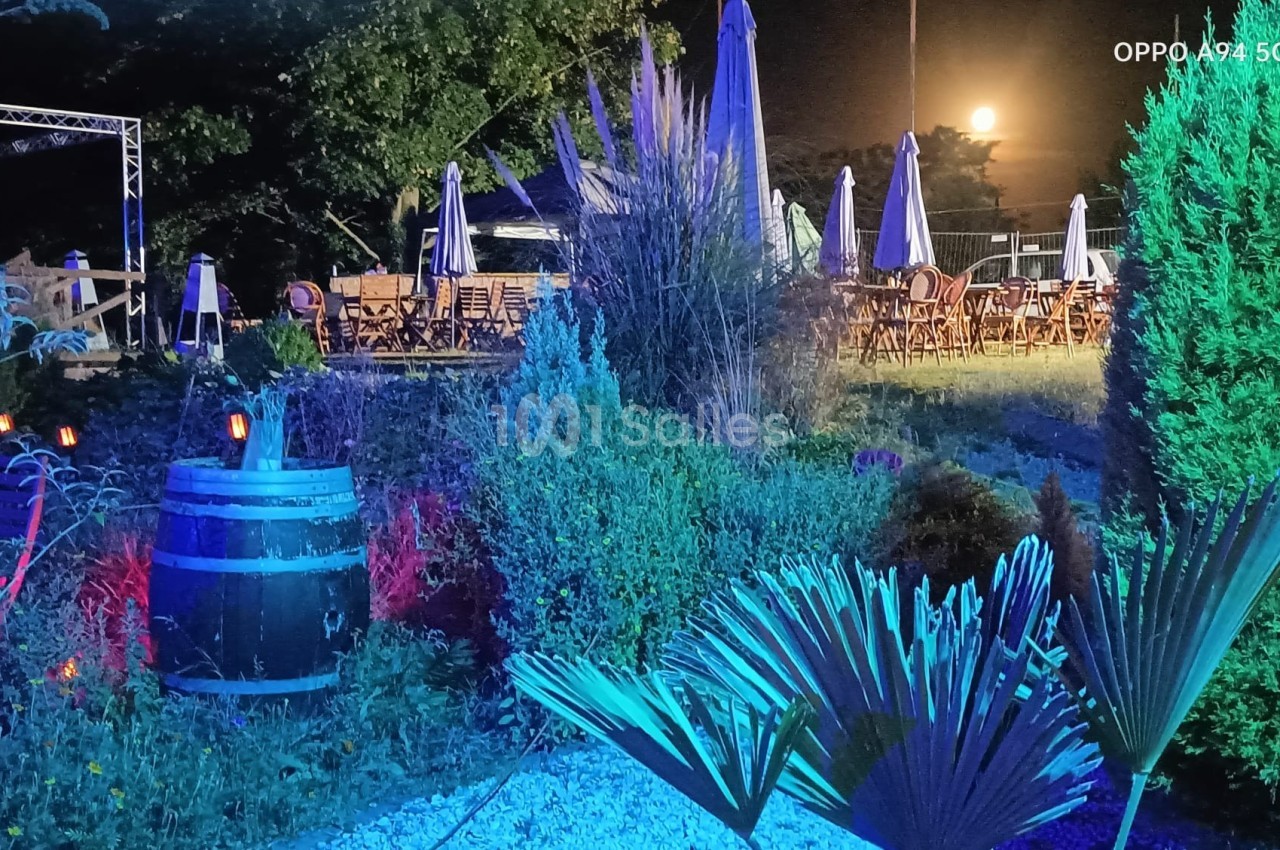 Jardin éclairé de nuit avec végétation, tonneau décoratif et terrasse en arrière-plan sous une pleine lune.