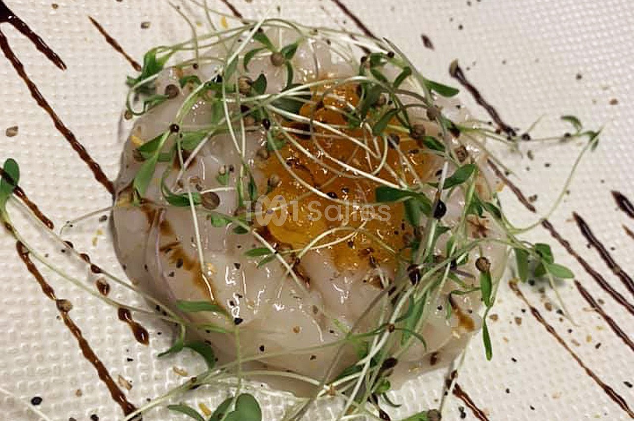 Tranche de carpaccio de Saint-Jacques garnie de pousses vertes, graines et filet de sauce sur assiette texturée.