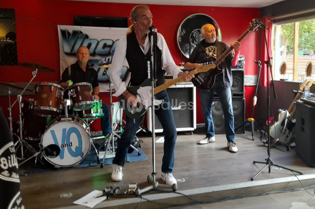Un groupe de musiciens joue sur une petite scène dans un bar, avec batterie, guitare et basse.