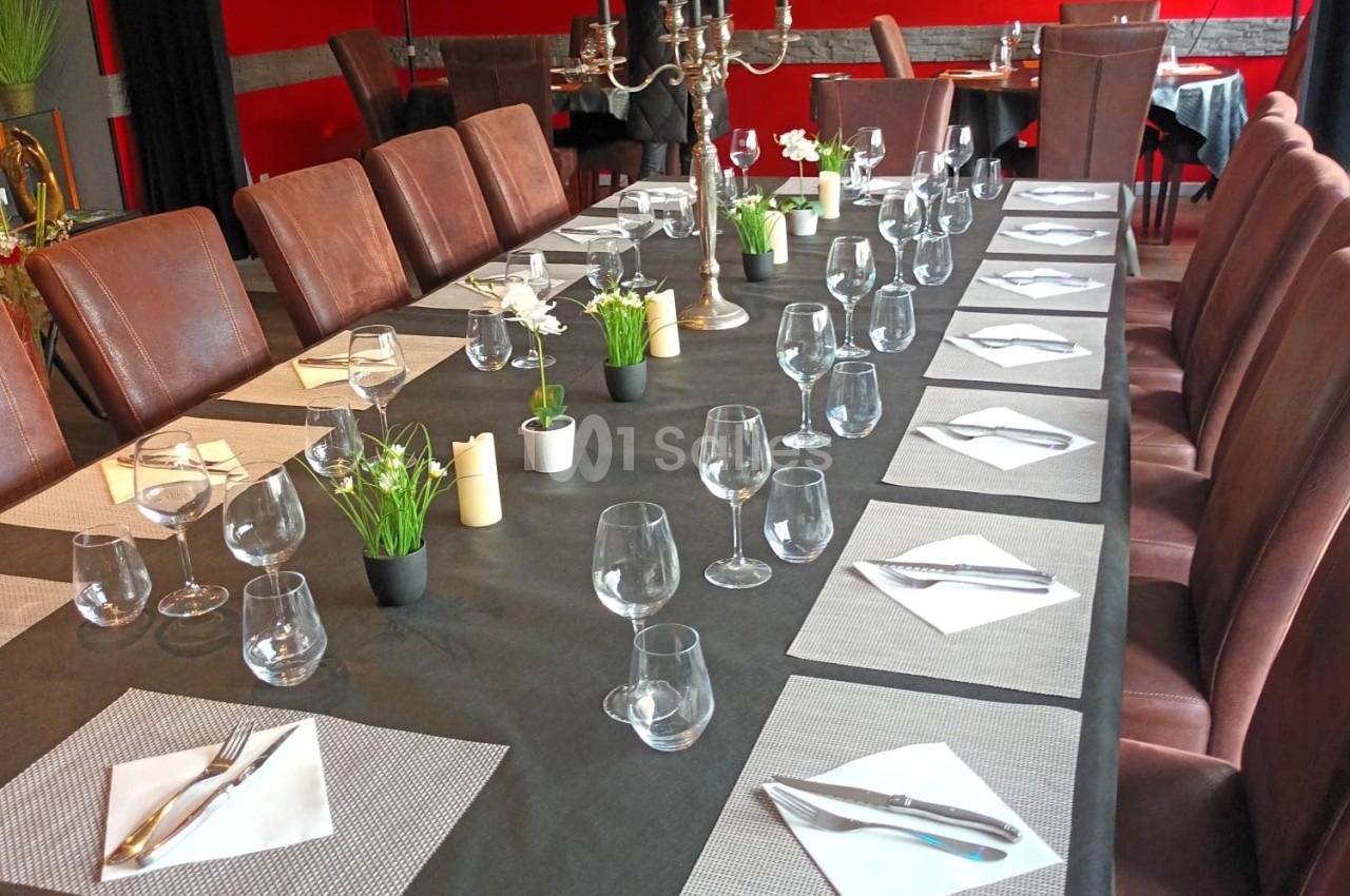 Grande table rectangulaire dressée avec nappes noires, verres, couverts et serviettes, dans une salle de restaurant rouge.
