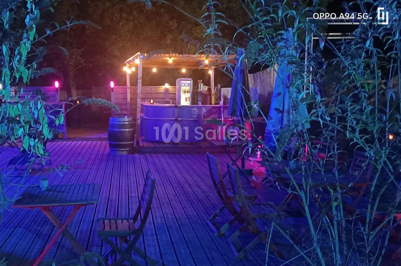 Terrasse en bois éclairée par des lumières colorées, avec un bar en arrière-plan et des tables vides au premier plan.
