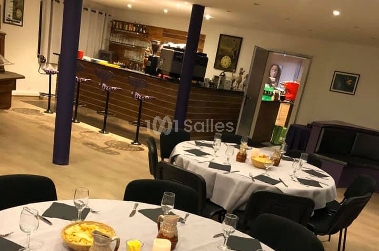Salle aménagée avec des tables rondes dressées, un bar en arrière-plan et un éclairage chaleureux.