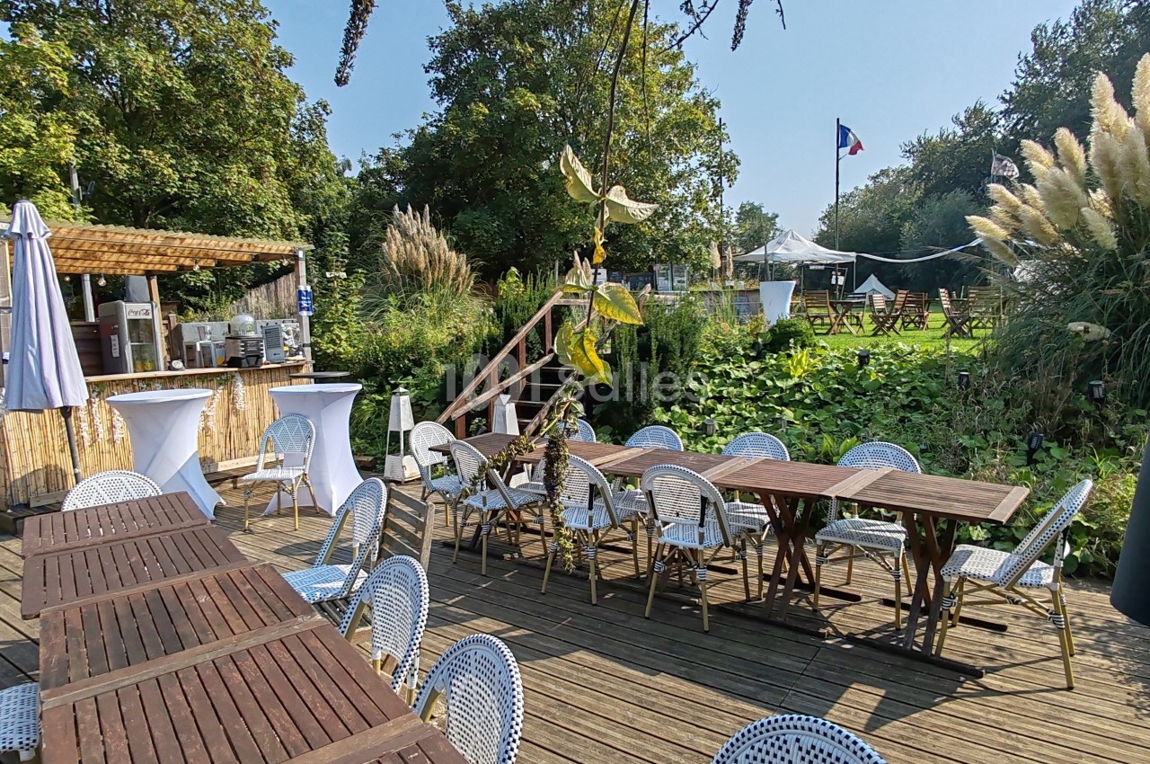 Terrasse en bois avec tables et chaises, entourée de végétation, sous un ciel dégagé.