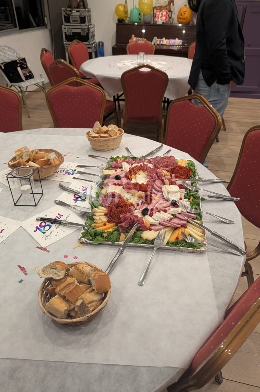 Plateau de charcuterie et fromages décoré avec fruits, entouré de couverts, sur une table dressée avec paniers de pain.