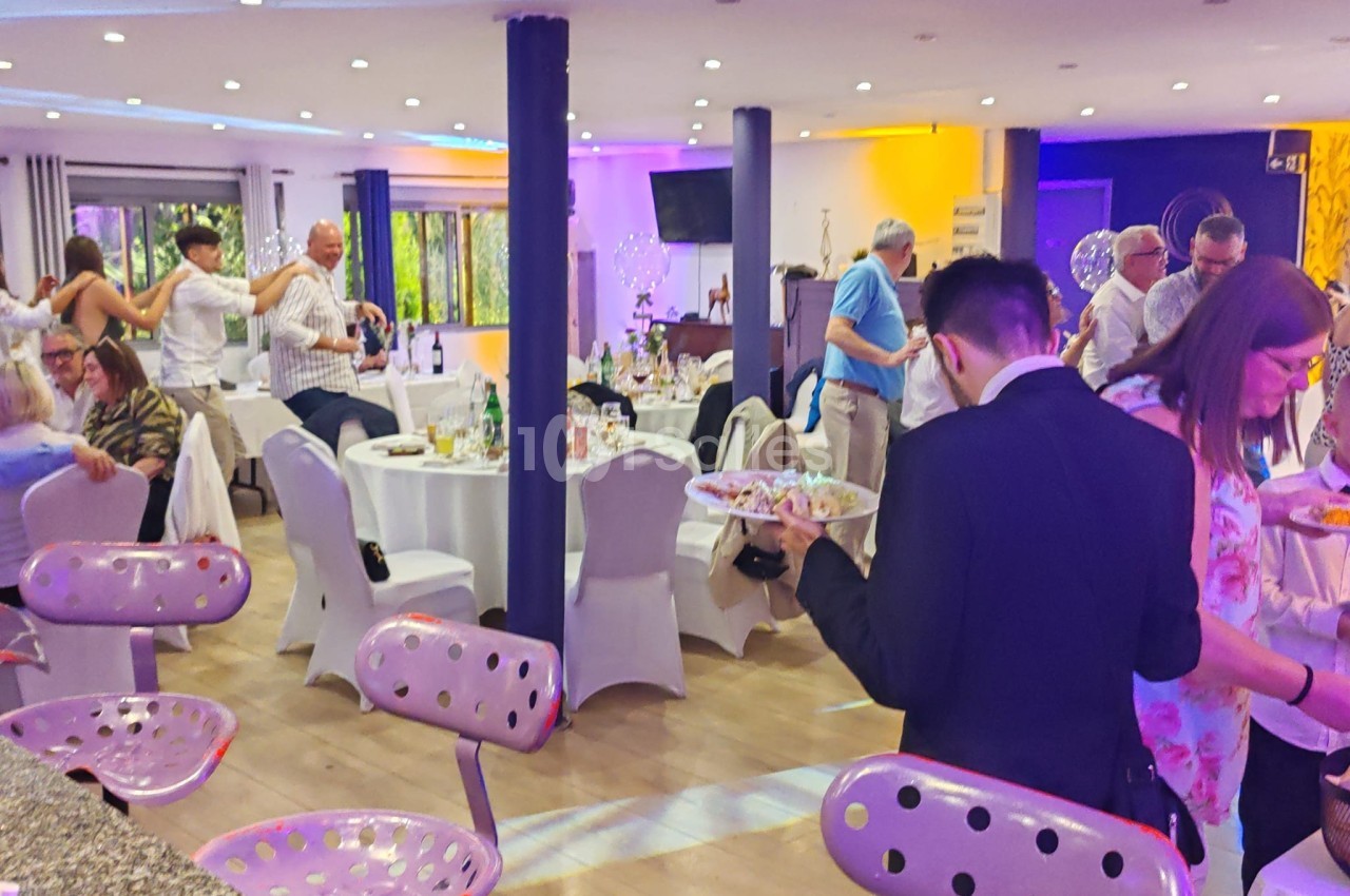 Salle de réception animée avec des invités debout et assis autour de tables décorées, éclairage coloré en arrière-plan.