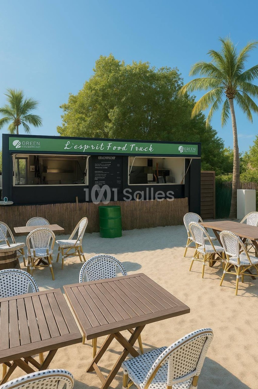 Food truck sur une plage avec des tables et chaises en rotin, entouré de palmiers et d'arbres sous un ciel dégagé.