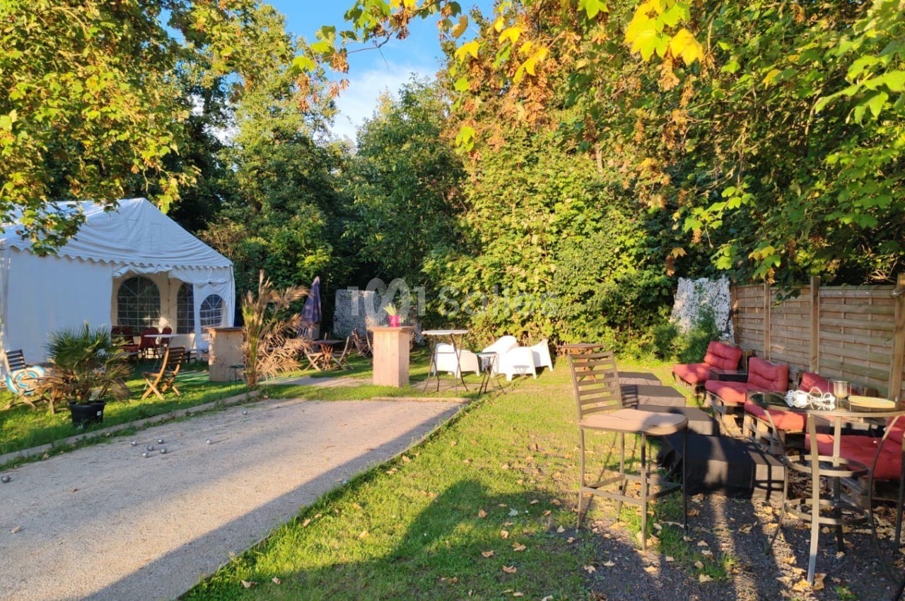 Espace extérieur avec tables, chaises et une tente blanche, entouré de verdure sous une lumière ensoleillée.