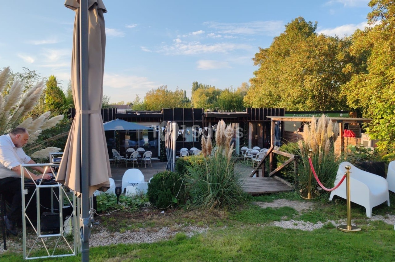 Un homme assis à une table en extérieur près d'une terrasse entourée de végétation et de mobilier de jardin.