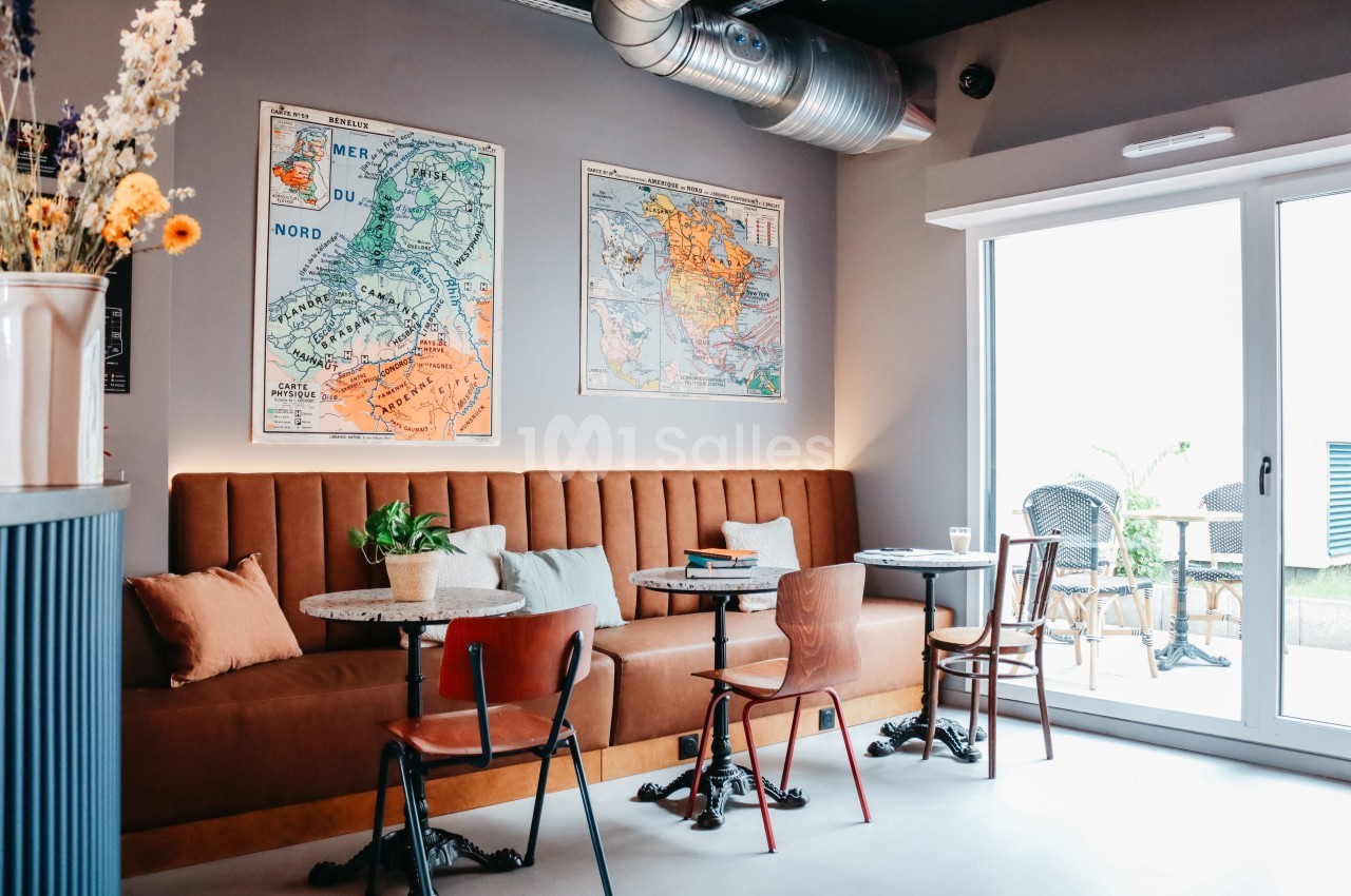 Banquette en cuir marron avec tables et chaises, décorée de cartes murales, près d'une baie vitrée donnant sur une terrasse.