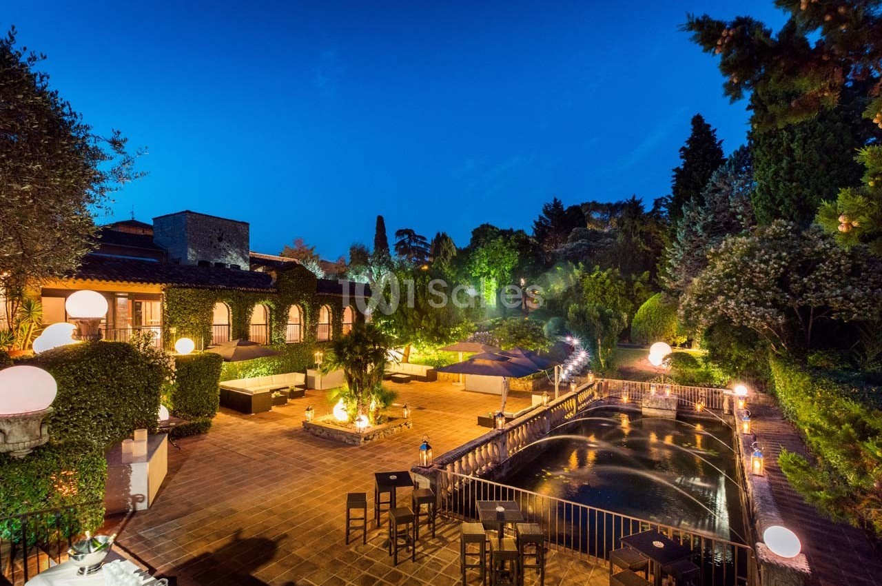 Terrasse illuminée en soirée avec tables, chaises, bassin d'eau et végétation luxuriante entourant l'espace.