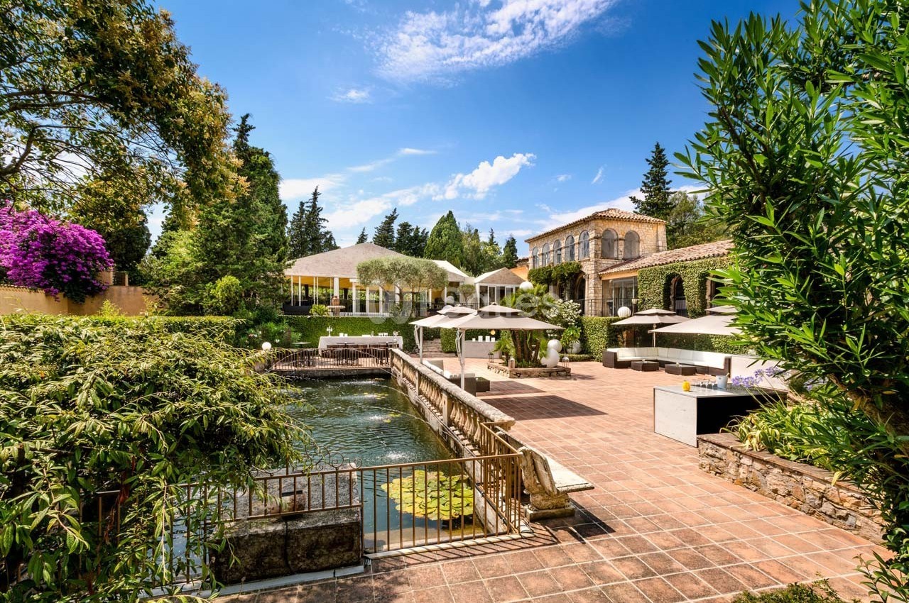 Jardin avec bassin, terrasse en pierre, parasols et bâtiments en pierre sous un ciel bleu ensoleillé.
