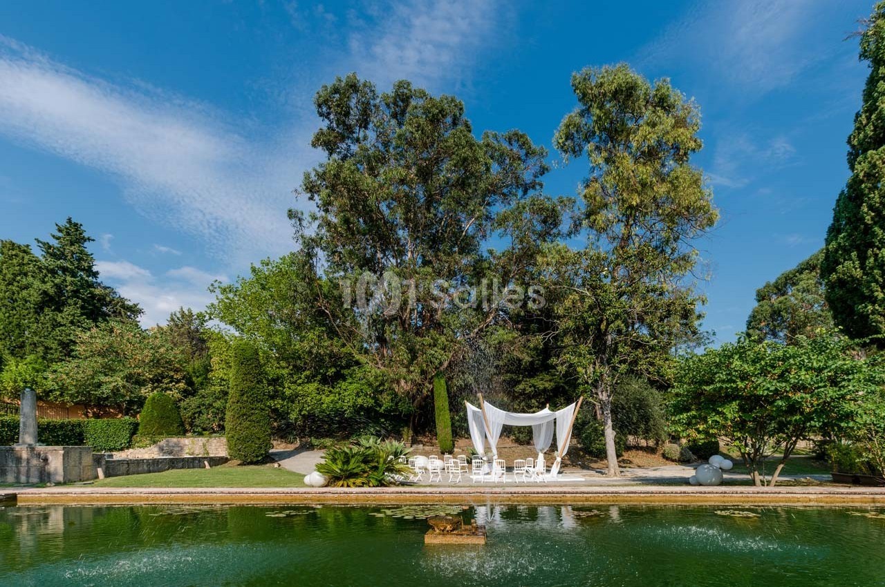 Un bassin entouré de verdure avec une arche blanche décorative et des chaises disposées sous des arbres.
