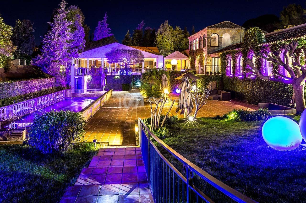 Jardin éclairé de nuit avec piscine, terrasse en pierre et bâtiments entourés de végétation.