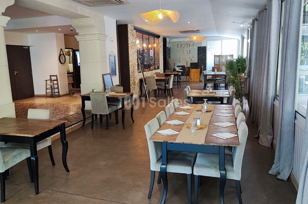 Salle de restaurant lumineuse avec tables dressées, chaises rembourrées et décor sobre mêlant bois et pierre.