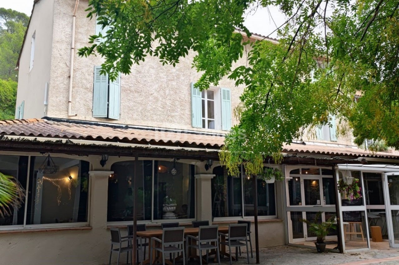 Façade d'une maison provençale avec volets verts, terrasse aménagée et arbres ombrageant l'entrée.