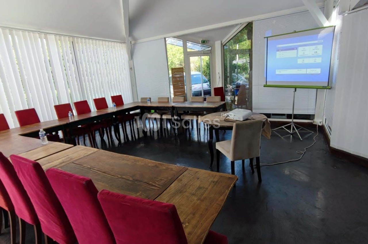 Salle de réunion lumineuse avec tables en bois disposées en U, chaises rouges et écran de projection allumé.