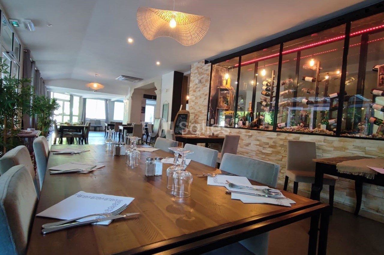 Salle de restaurant lumineuse avec tables dressées, décor moderne et mur vitré exposant des bouteilles de vin.