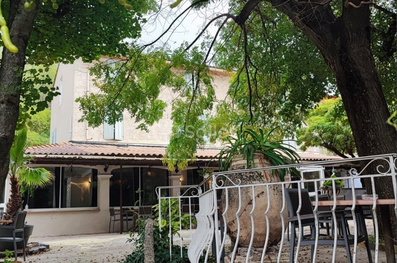 Maison en pierre entourée d'arbres avec une terrasse aménagée et une grande jarre décorative au premier plan.