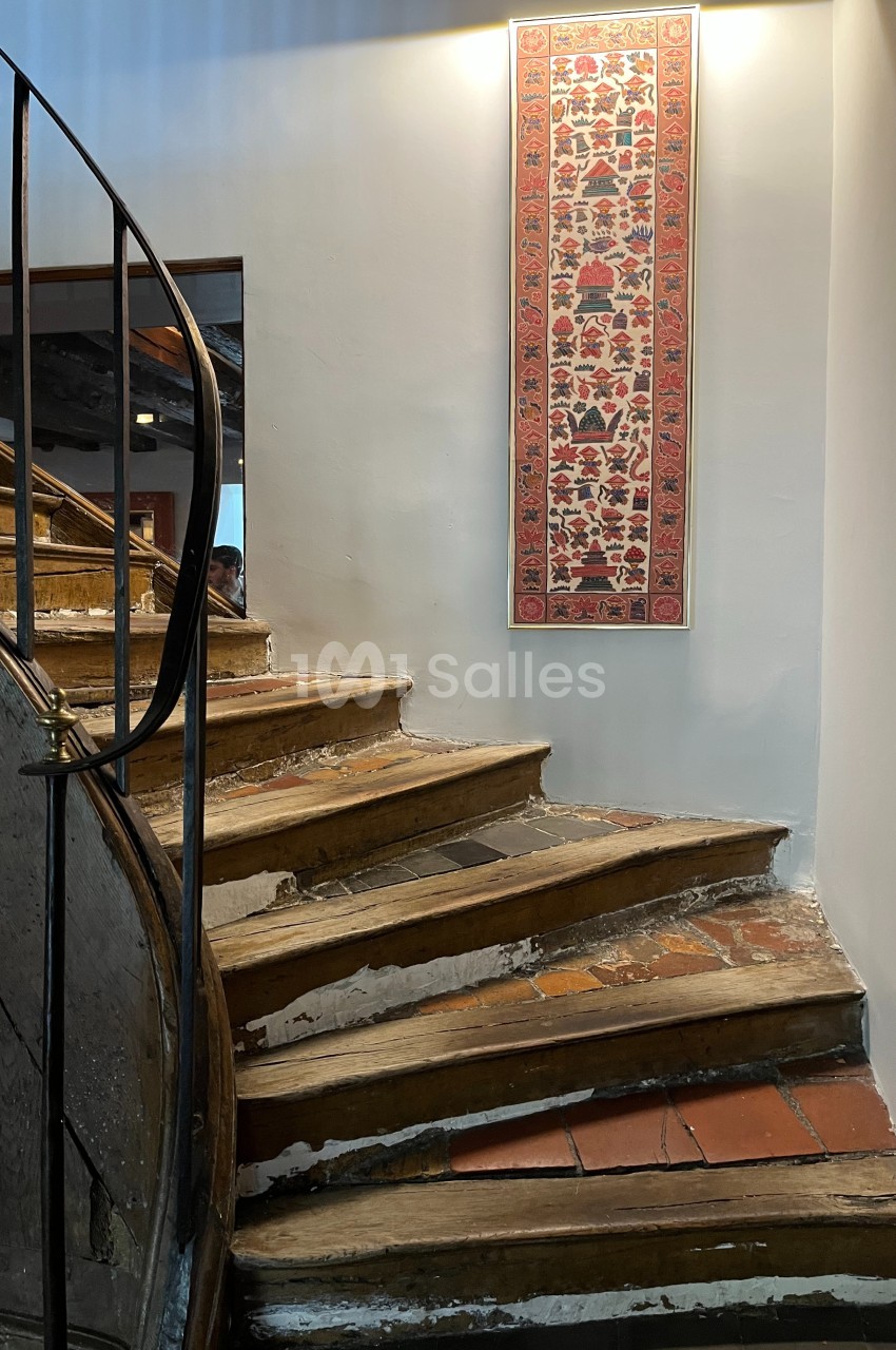 Escalier en bois usé avec une rampe en métal, éclairé par une applique murale, et décoré d'un tapis mural vertical.