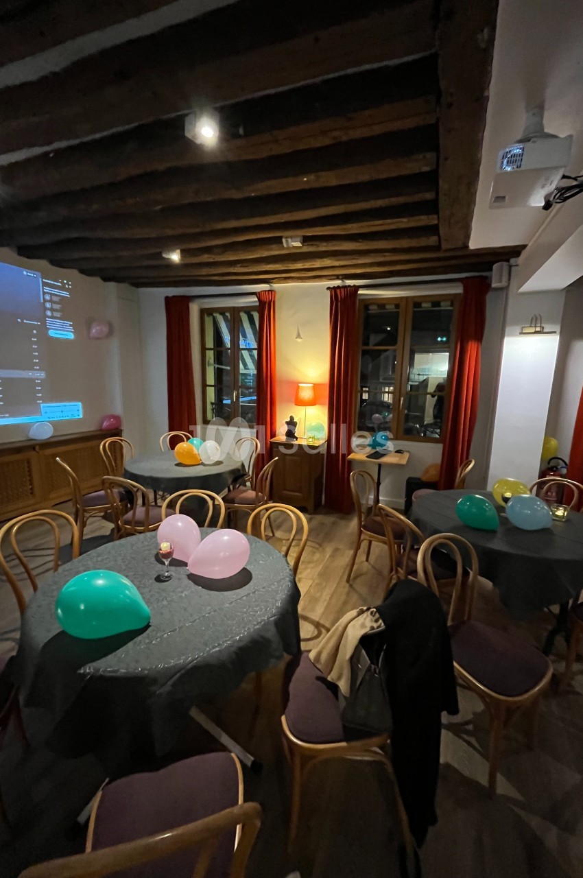 Salle décorée pour un événement avec des tables rondes, des ballons colorés et un éclairage chaleureux.