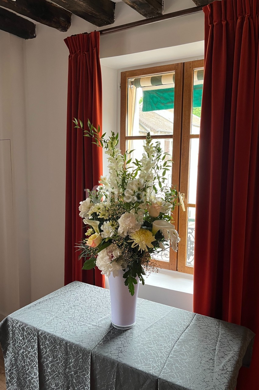 Bouquet de fleurs blanches et jaunes dans un vase blanc, posé sur une table devant une fenêtre encadrée de rideaux rouges.