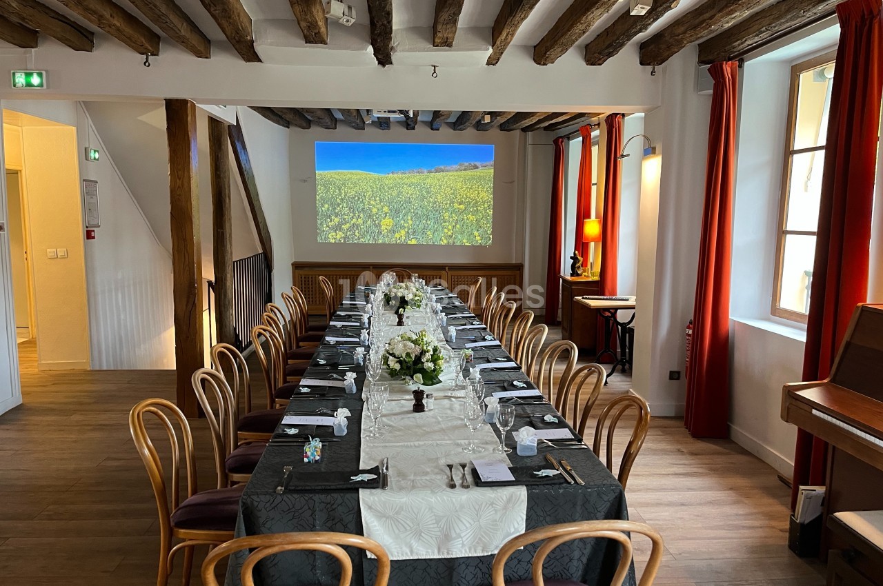 Salle de réception avec une longue table dressée, décorée de fleurs blanches, sous un plafond en poutres apparentes.
