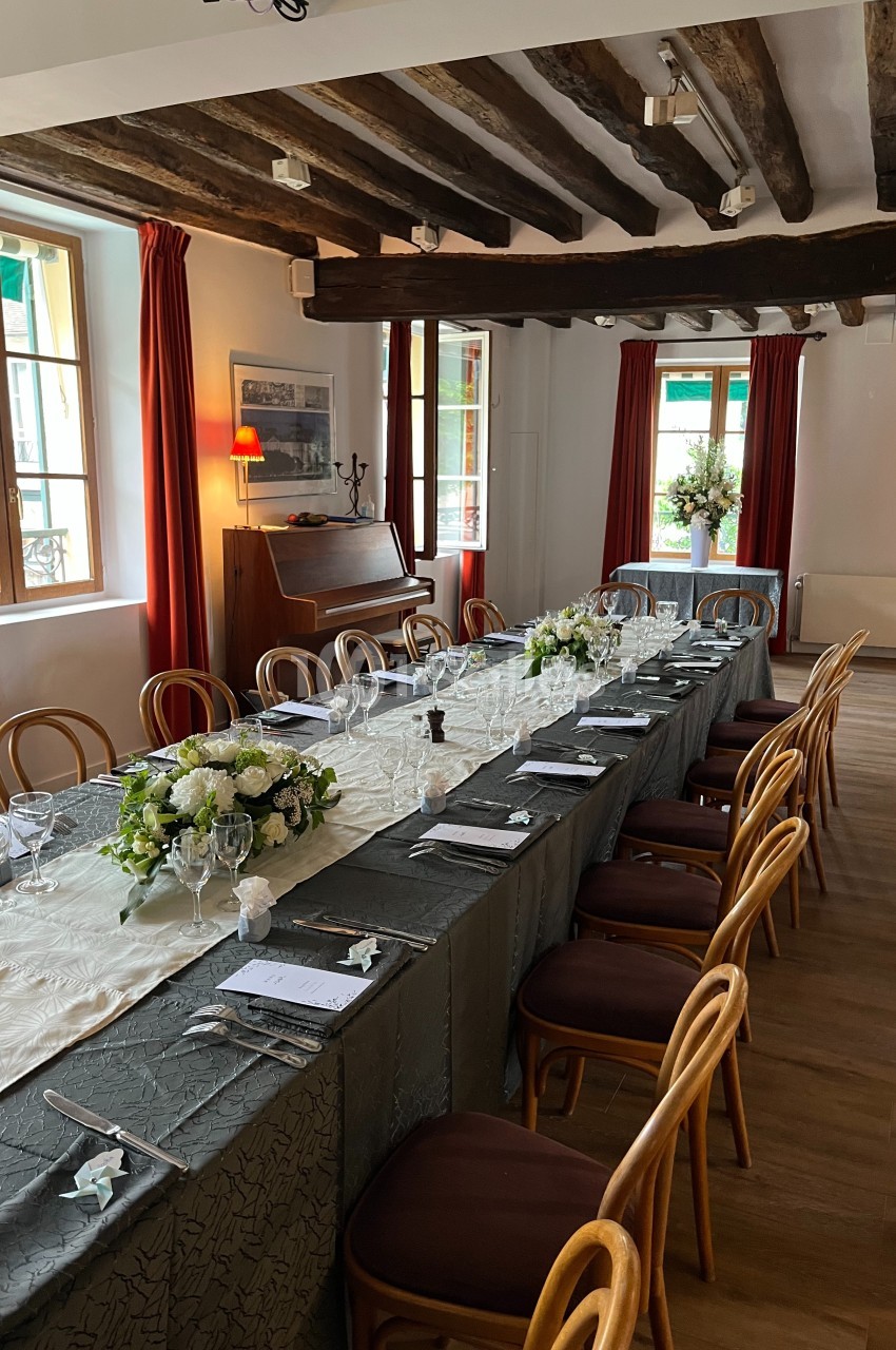 Table dressée dans une salle lumineuse avec poutres apparentes, nappes élégantes, fleurs et chaises en bois.