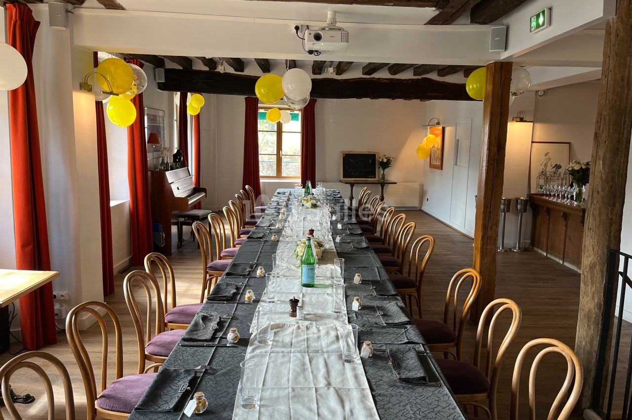 Salle décorée avec une longue table dressée, chaises en bois, ballons jaunes et lumière naturelle entrant par les fenêtres.