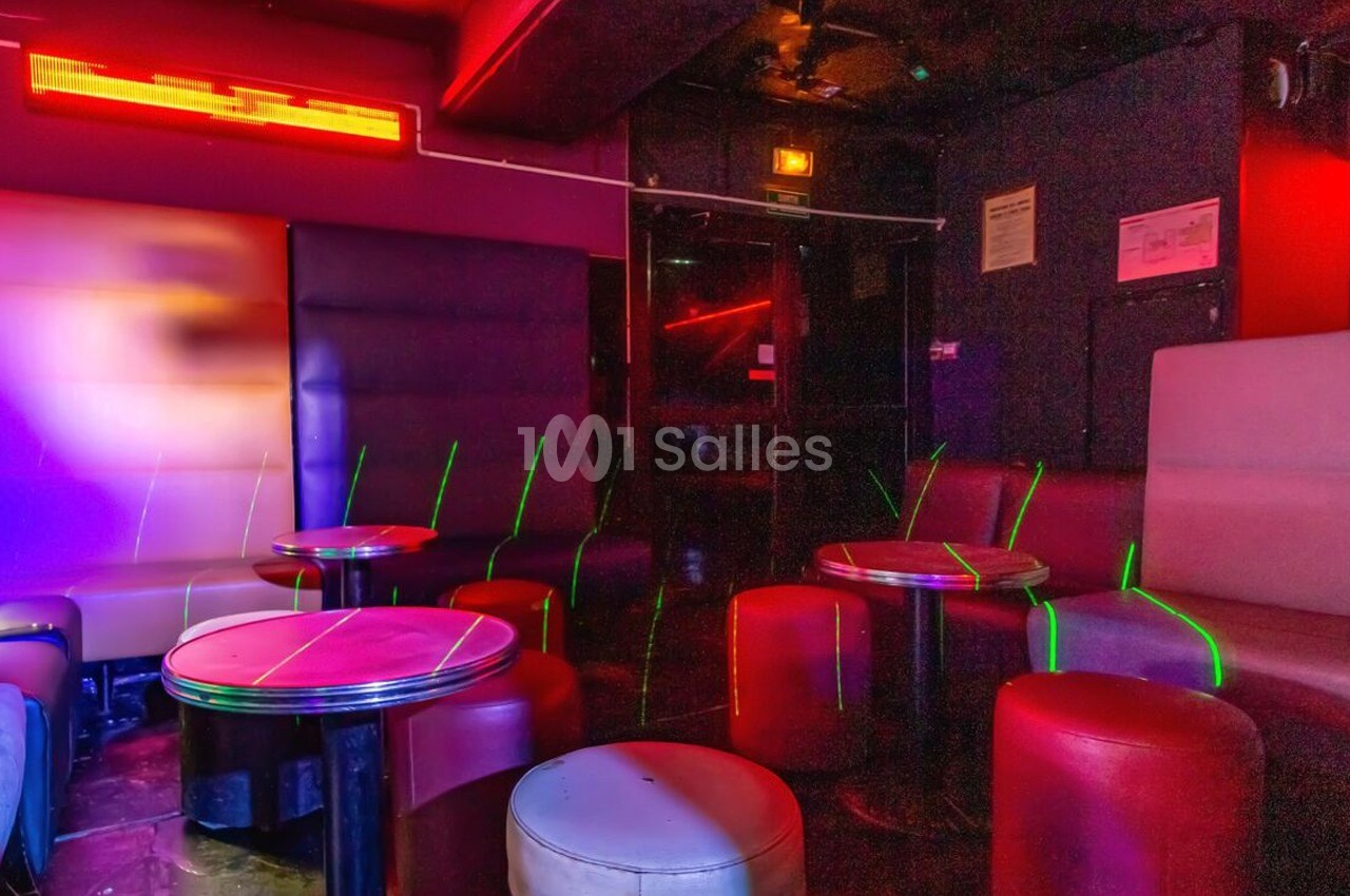 Salle de bar avec banquettes, tables rondes et éclairage tamisé aux tons rouges et violets.
